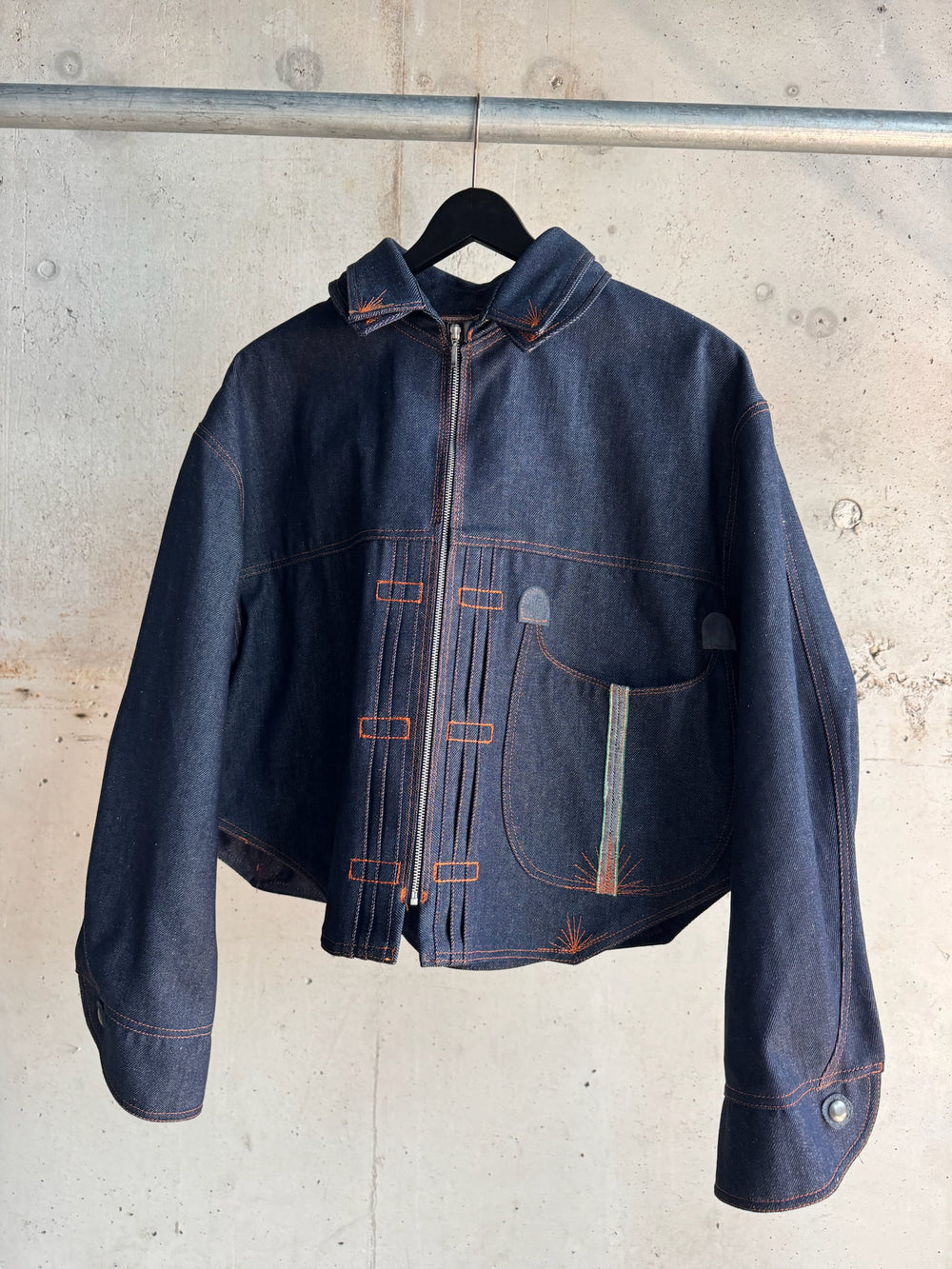 Dark indigo denim jacket with orange contrast stitching, asymmetrical front zipper, and curved oversized pocket, hanging on a black hanger against a raw concrete wall.