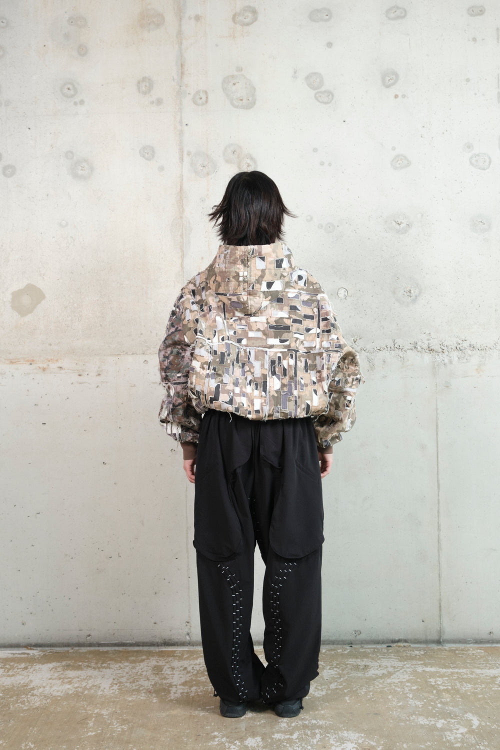 A model stands with their back to the camera against a minimalist concrete wall, wearing a distressed patchwork hoodie with an earthy camouflage pattern. The hoodie features frayed textures, layered fabric panels, and a structured hood. Paired with black wide-leg pants that have draped fabric details and silver contrast stitching, the outfit blends deconstructed aesthetics with a utilitarian edge.