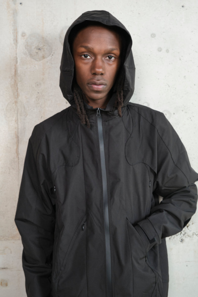 Male Model with black technical jacket. grey concrete wall. One hand in pocket. Technical lines in the design of the jacket.