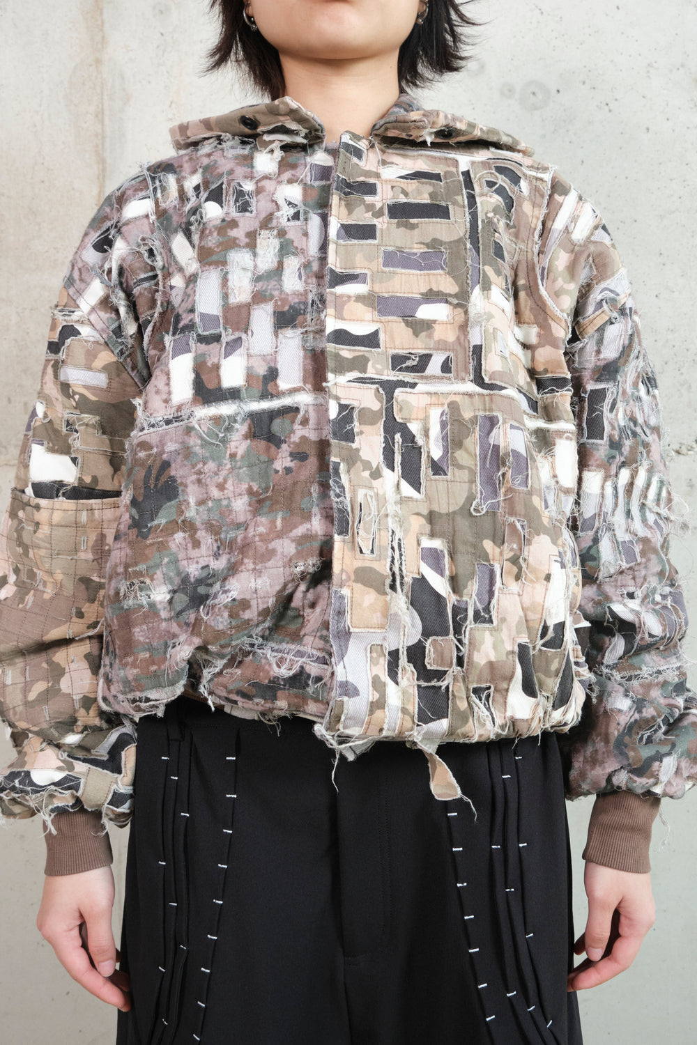 Close-up of a model wearing a distressed patchwork jacket with a camouflage pattern. The jacket features frayed edges, layered fabric textures, and a deconstructed aesthetic. The model pairs it with black pants adorned with white contrast stitching. The combination of raw, unfinished details and structured elements creates a bold, avant-garde look.