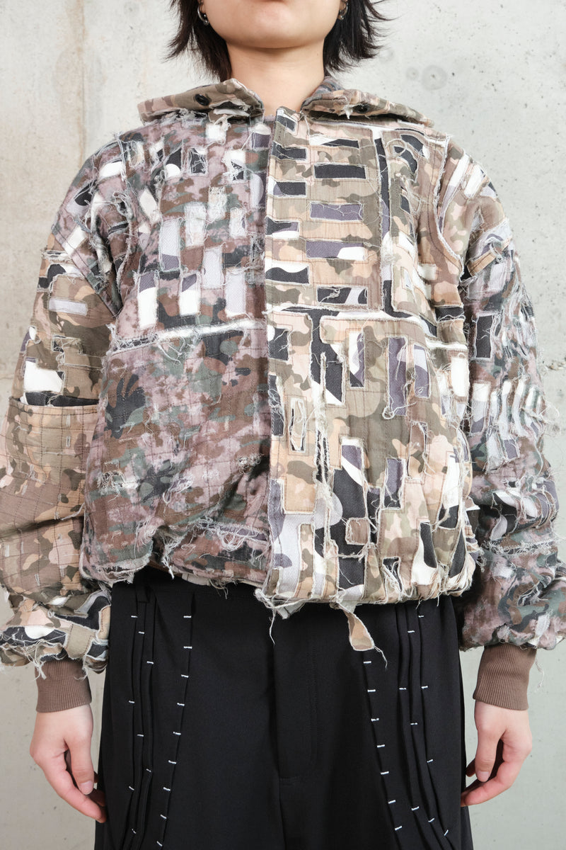 Close-up of a model wearing a distressed patchwork jacket with a camouflage pattern. The jacket features frayed edges, layered fabric textures, and a deconstructed aesthetic. The model pairs it with black pants adorned with white contrast stitching. The combination of raw, unfinished details and structured elements creates a bold, avant-garde look.