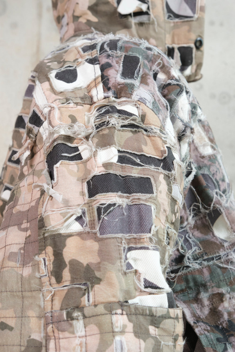 Close-up of a distressed patchwork hoodie with an earthy camouflage pattern. The fabric features frayed cut-out patches and layered stitching, creating a deconstructed, textured look. The detailed craftsmanship emphasizes the raw, rugged aesthetic of the garment.