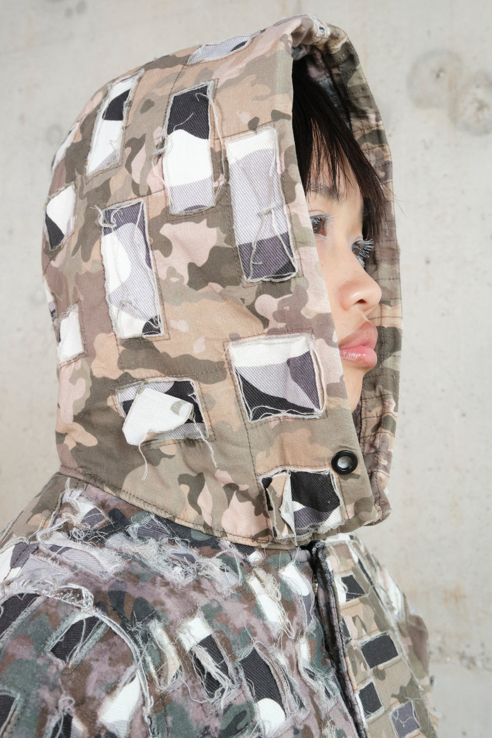 Close-up of a model wearing a distressed patchwork hoodie with an earthy camouflage pattern. The hood features cut-out fabric patches with frayed edges, adding texture and depth to the design. The model’s face is partially visible, highlighting their soft features and white mascara on the eyelashes. The composition emphasizes the raw, deconstructed aesthetic of the garment.