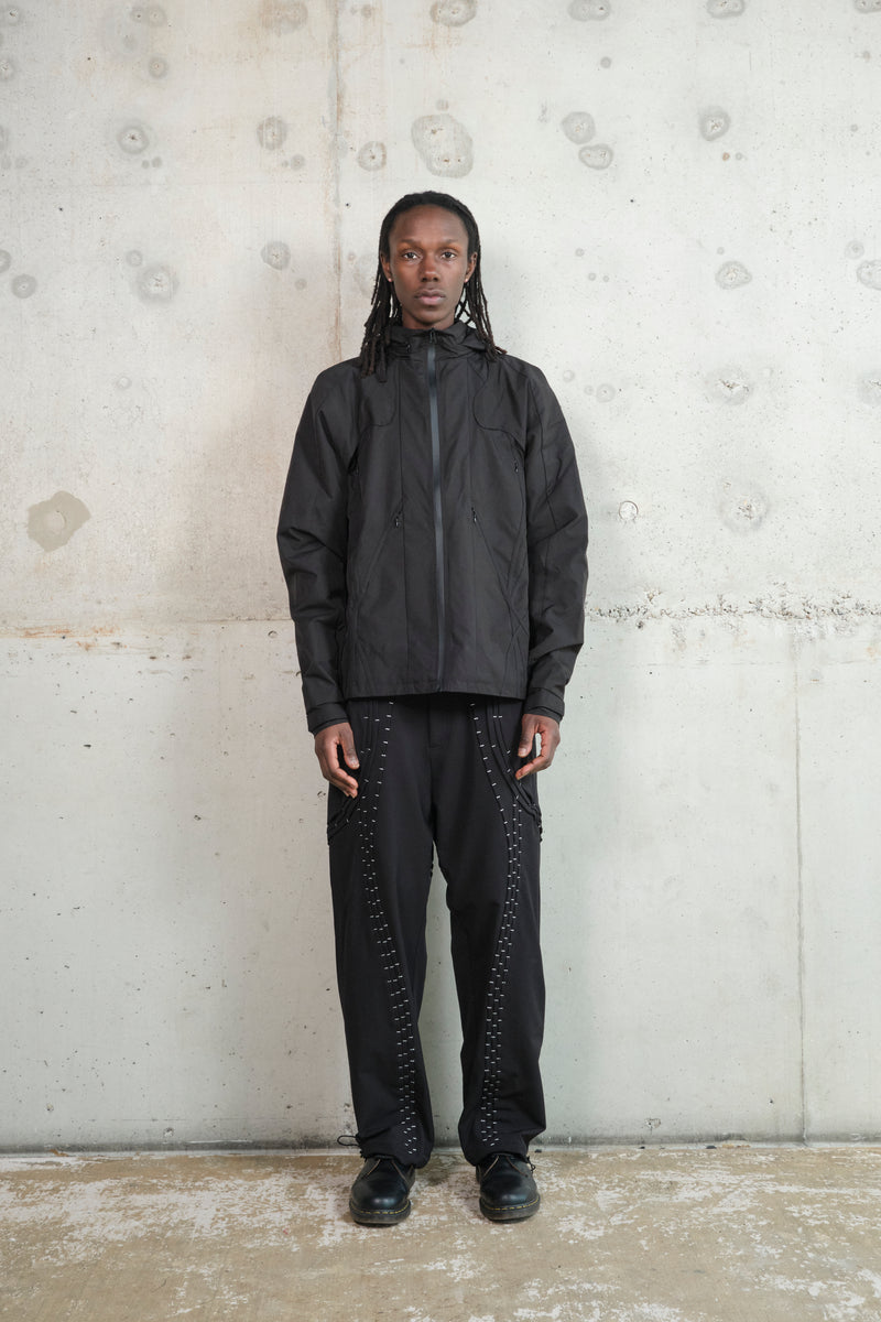 A man with dark skin and long dreadlocks stands upright against a minimalist concrete wall. He is wearing a black technical jacket with a high zipper closure and zippered pockets. His black pants feature striking silver stitching in a curved pattern. He pairs the outfit with black leather shoes and maintains a calm, neutral facial expression. The industrial background and minimalist styling highlight the modern, streetwear-inspired aesthetic of the outfit.
