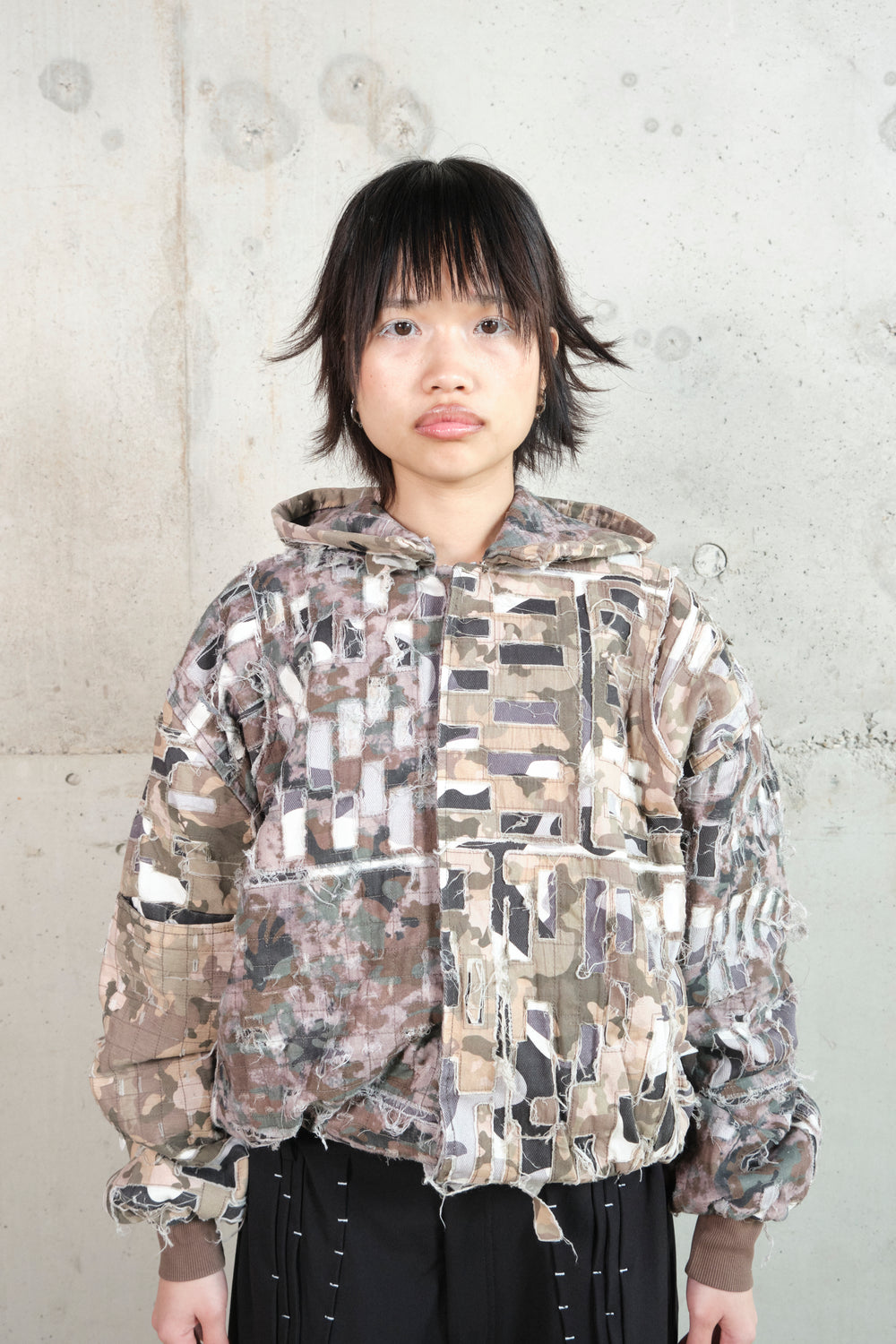 A model with short, tousled black hair stands against a minimalist concrete wall, wearing a distressed patchwork hoodie in earthy camouflage tones. The hoodie features frayed edges, layered fabric details, and an asymmetrical design. The model pairs it with black pants accented with white contrast stitching. Their neutral expression and relaxed stance emphasize the rugged, deconstructed aesthetic of the outfit.