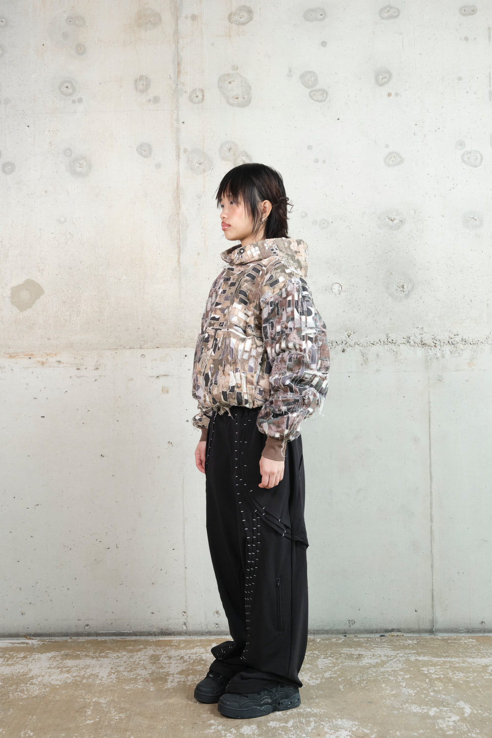A model with short black hair stands in profile against a minimalist concrete wall, wearing a distressed patchwork hoodie with an earthy camouflage pattern. The hoodie features frayed textures and a structured hood. Paired with black wide-leg pants accented with white contrast stitching and black sneakers, the outfit blends deconstructed and utilitarian aesthetics. The relaxed posture highlights the unique layering and details of the garments.