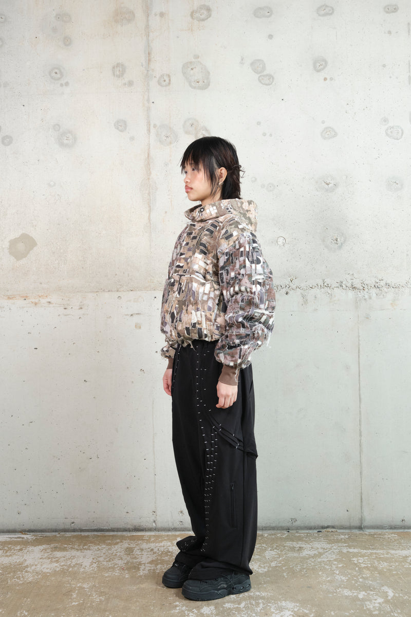 A model with short black hair stands in profile against a minimalist concrete wall, wearing a distressed patchwork hoodie with an earthy camouflage pattern. The hoodie features frayed textures and a structured hood. Paired with black wide-leg pants accented with white contrast stitching and black sneakers, the outfit blends deconstructed and utilitarian aesthetics. The relaxed posture highlights the unique layering and details of the garments.