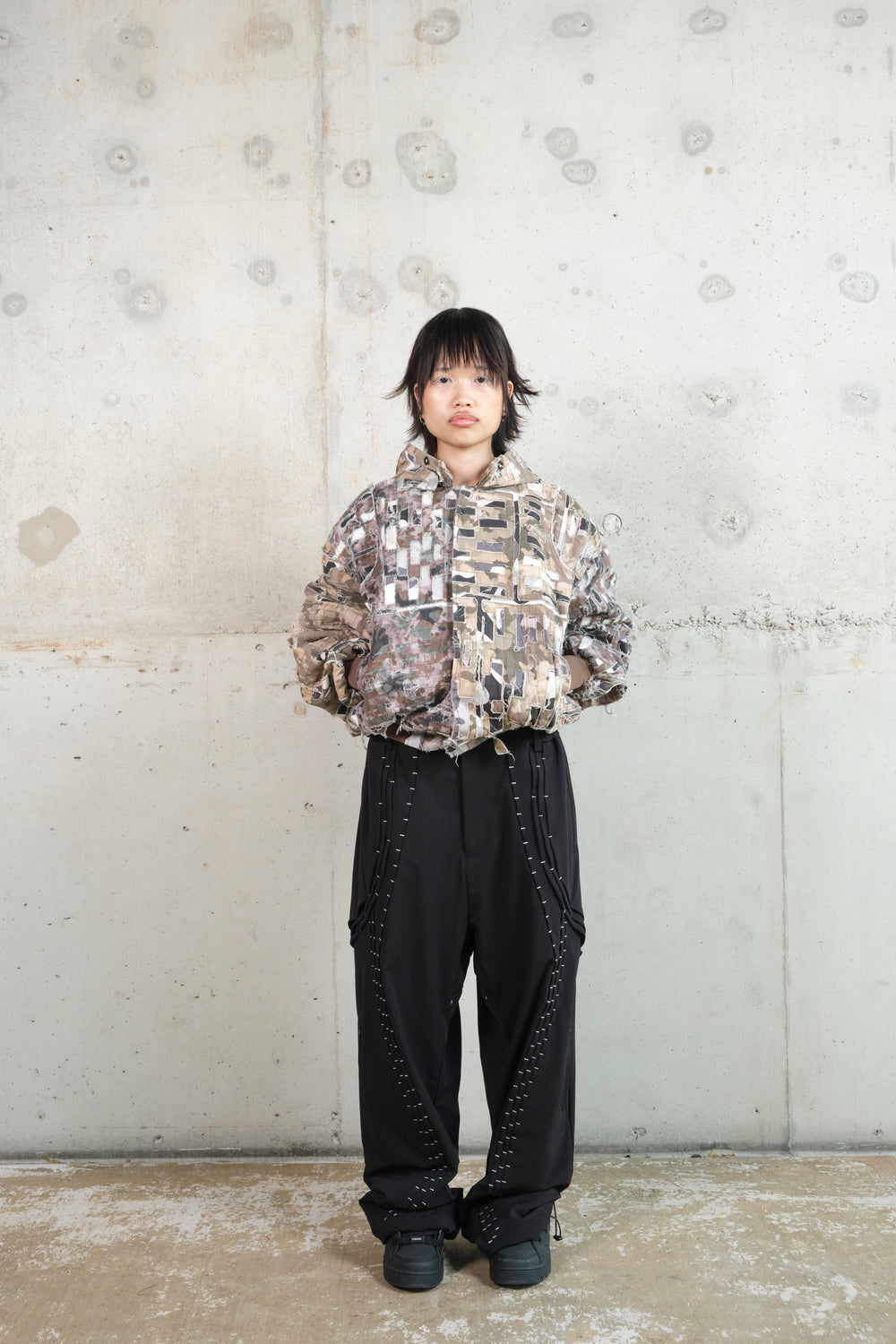 A model with short black hair stands against a minimalist concrete wall, wearing a distressed patchwork hoodie in earthy camouflage tones. The hoodie features frayed edges, layered fabric details, and an oversized fit. They pair it with black wide-leg pants adorned with intricate white stitching and black sneakers. Their stance is relaxed, highlighting the deconstructed, avant-garde aesthetic of the outfit.