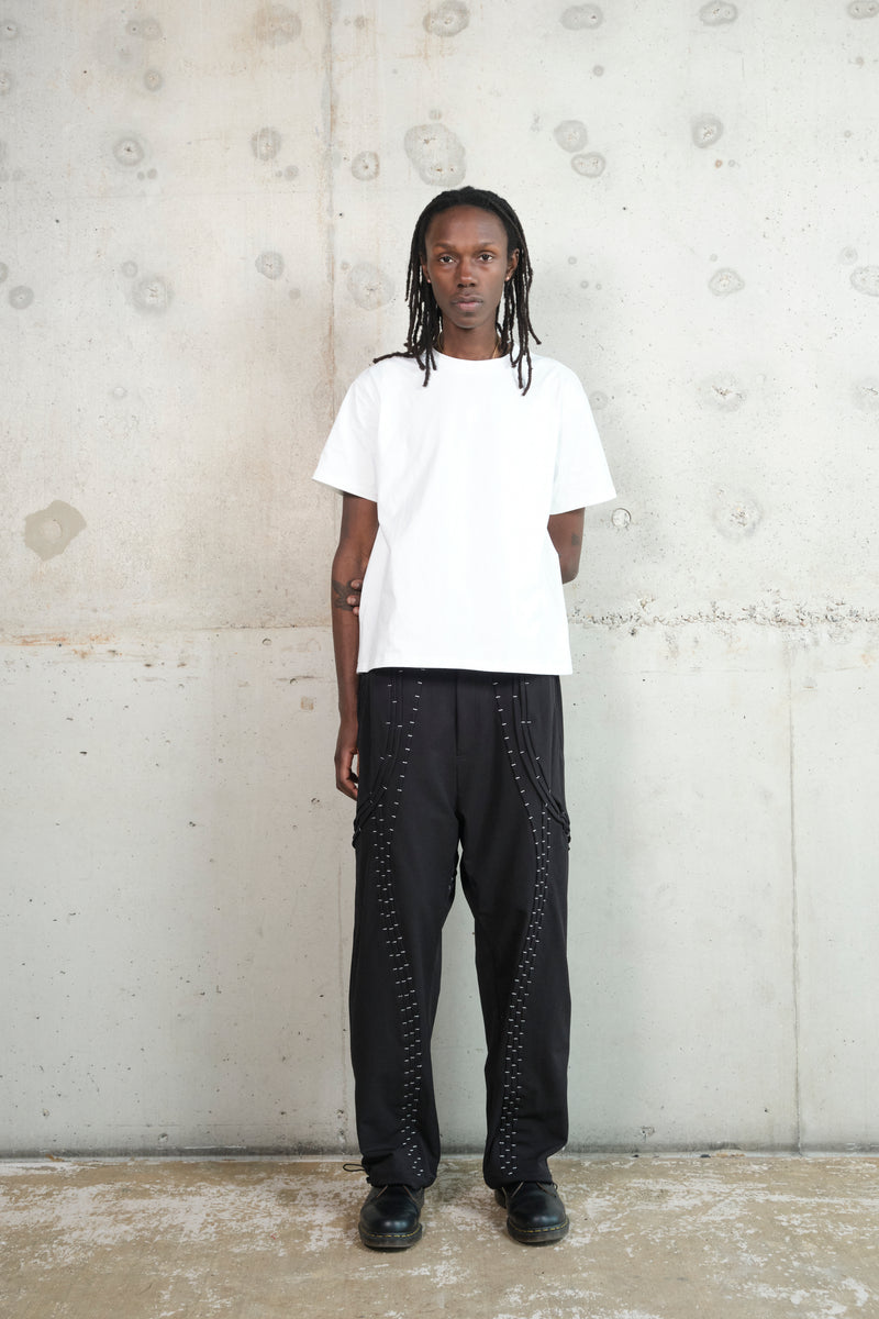 A male model with dark skin and long dreadlocks stands against a minimalist concrete wall. He is wearing a crisp white oversized T-shirt paired with black wide-leg pants featuring intricate silver stitching details. His black leather shoes complete the monochrome look. His neutral expression and relaxed stance emphasize the clean, modern aesthetic of the outfit.