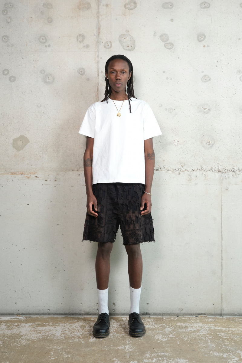 Model wearing a minimalist outfit featuring a plain white T-shirt, textured black shorts with a deconstructed patchwork design, white socks, and black leather shoes. The look is completed with a gold pendant necklace, standing against a raw concrete wall.