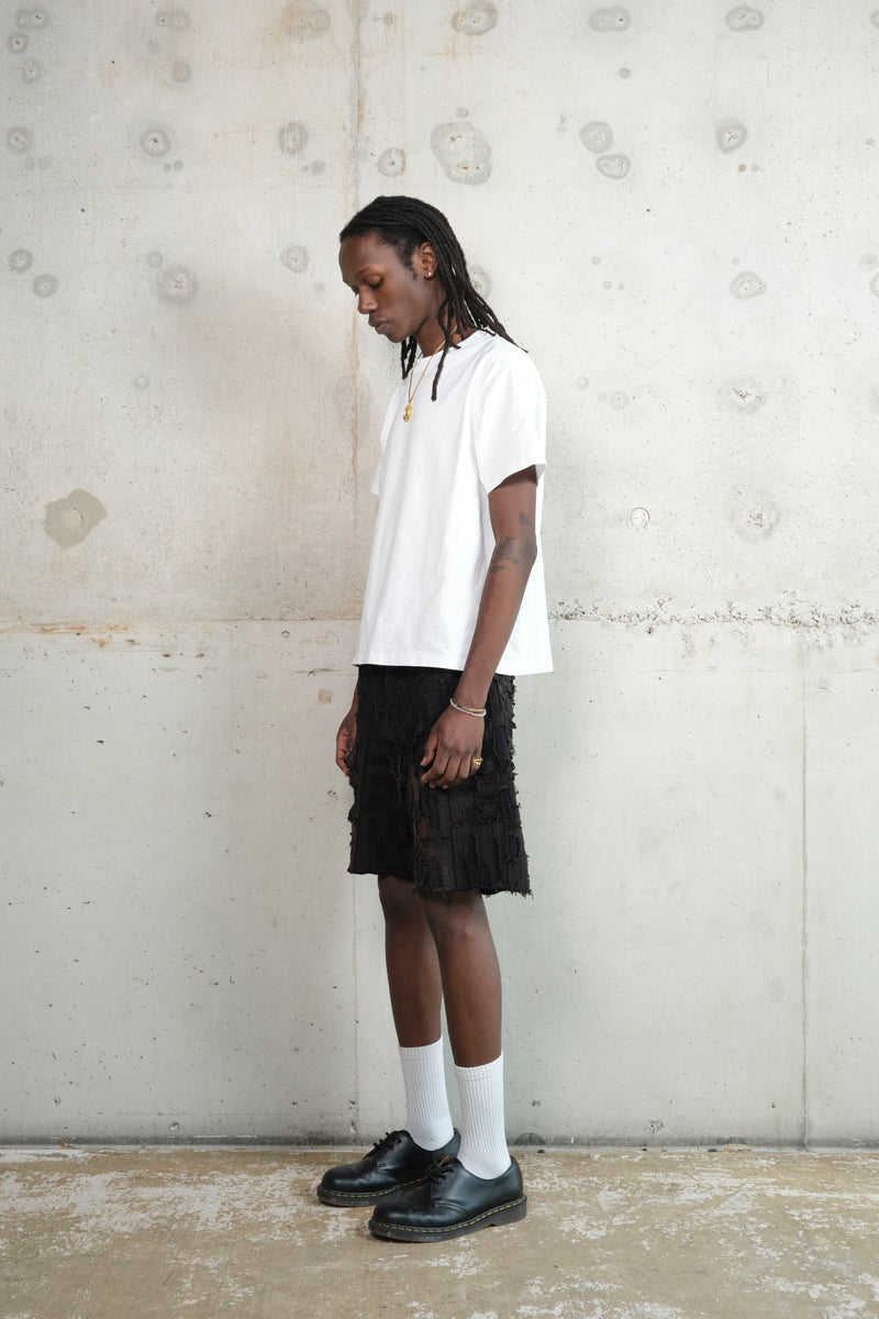 Model in a relaxed pose, wearing a minimalist outfit featuring a white oversized T-shirt, black textured shorts with a deconstructed design, white socks, and black leather shoes. Accessories include a gold pendant necklace and rings. The model stands against a raw concrete wall.