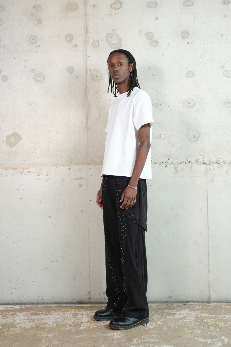 A male model with dark skin and long dreadlocks stands in a relaxed pose against a textured concrete wall. He wears a white oversized T-shirt paired with black wide-leg pants featuring silver stitching details. His black leather shoes complete the monochrome ensemble. The side profile highlights the clean lines and minimalistic aesthetic of the outfit.