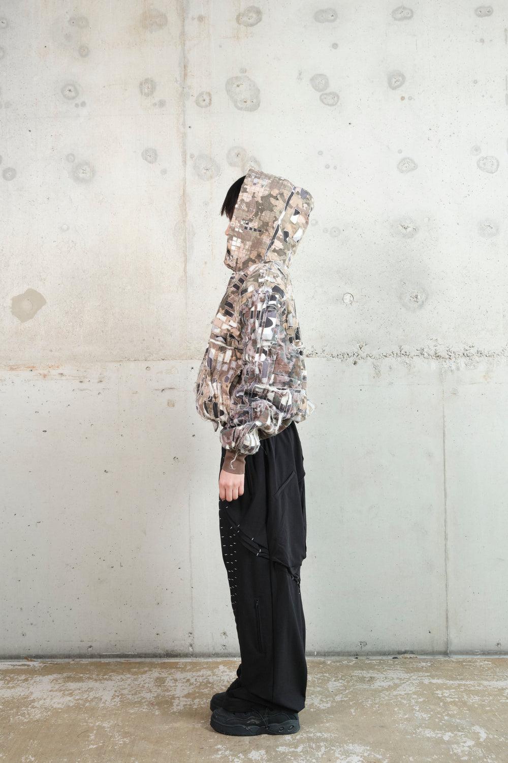 A model stands in profile against a minimalist concrete wall, wearing a distressed patchwork hoodie with an earthy camouflage pattern. The hood is up, partially covering their face, emphasizing the deconstructed and layered fabric details. Paired with black wide-leg pants featuring structured paneling and silver stitching, the outfit blends rugged textures with a modern, utilitarian silhouette.