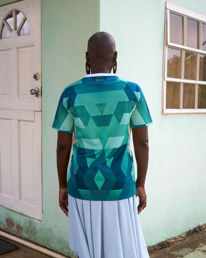 A woman with a shaved head and large gold earrings stands with her back to the camera in front of a pastel green house with a white door and a window. She wears a green and white patterned sports jersey with geometric shapes on the back and a flowing light blue skirt. The setting is a residential area with a textured wall and a simple, natural background