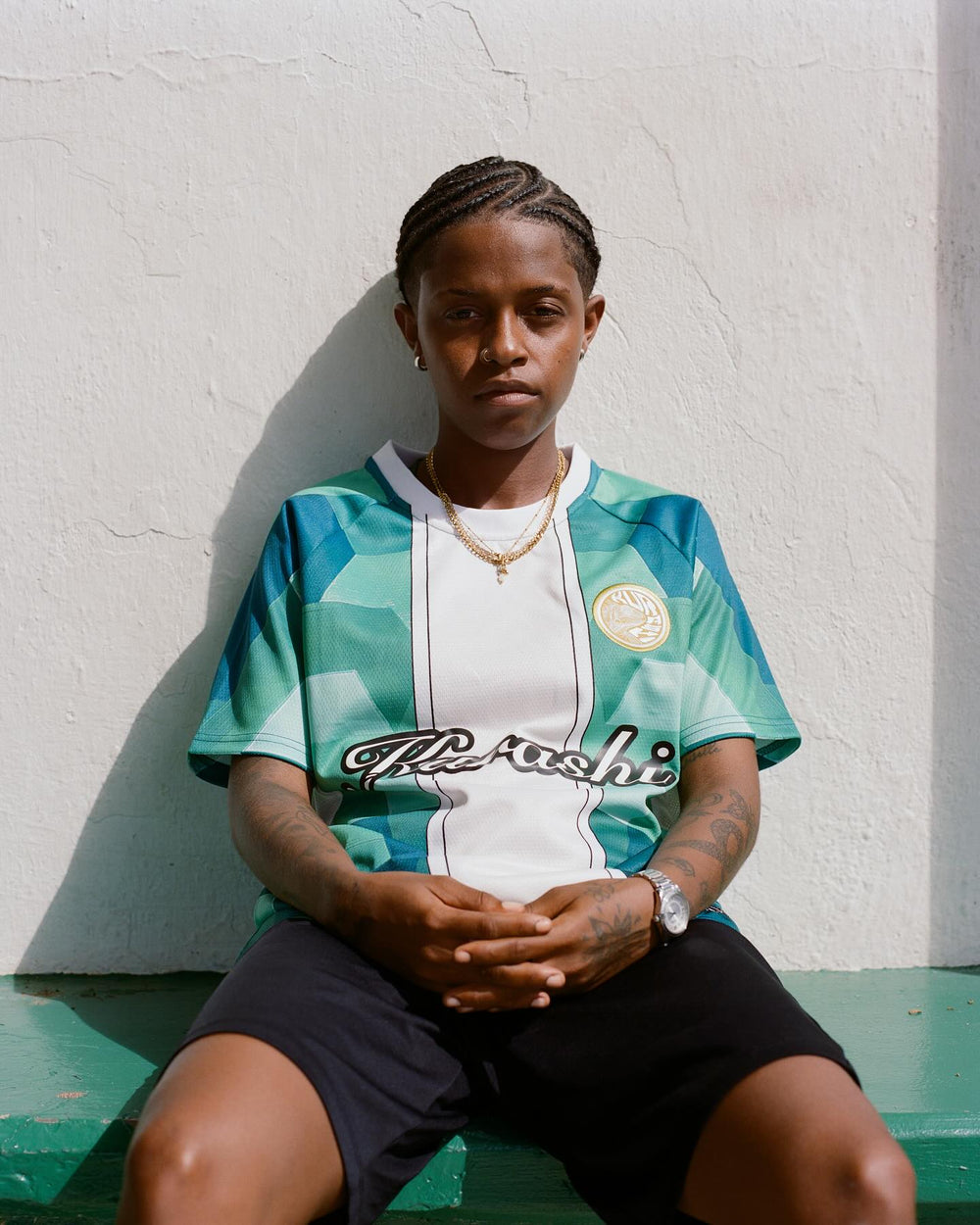 A person with neatly braided hair sits against a textured white wall, looking directly at the camera with a calm and confident expression. They are wearing a green and white geometric-patterned sports jersey with the word ‘Kurashi’ written in black cursive across the chest, along with a gold emblem on the upper left side. They also wear black shorts, a gold chain necklace, a silver watch, and have tattoos on their arms. Their hands are resting together in a relaxed posture, and they are seated on a green le