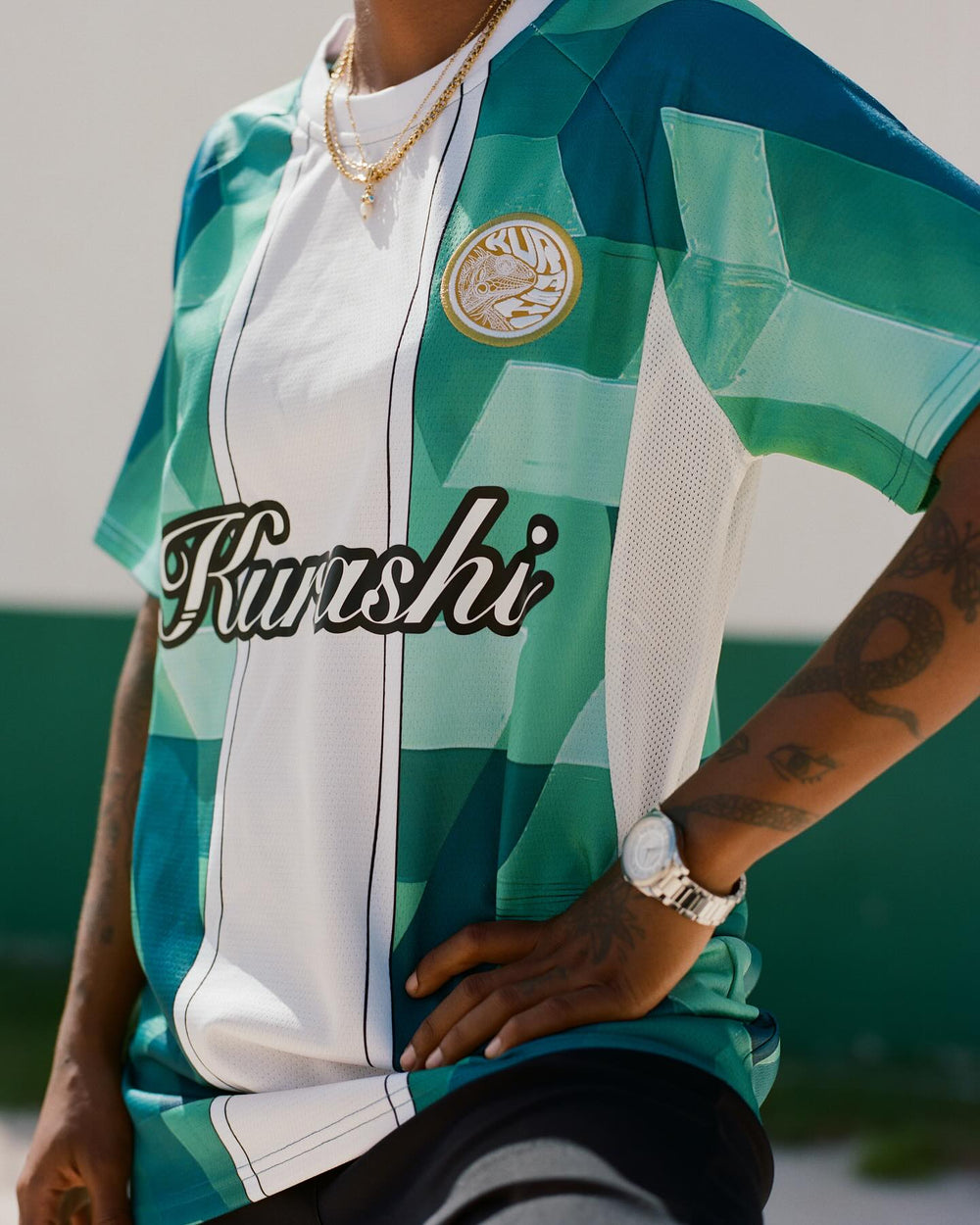 A close-up of a person wearing a green and white geometric-patterned sports jersey with the word ‘Kurashi’ written in black cursive across the chest. A gold emblem with a circular design is on the upper left side of the jersey. The person has tattoos on their arms, including an eye and a snake, and is wearing a silver watch and layered gold necklaces. They are posing with one hand on their hip, with a blurred green and white background