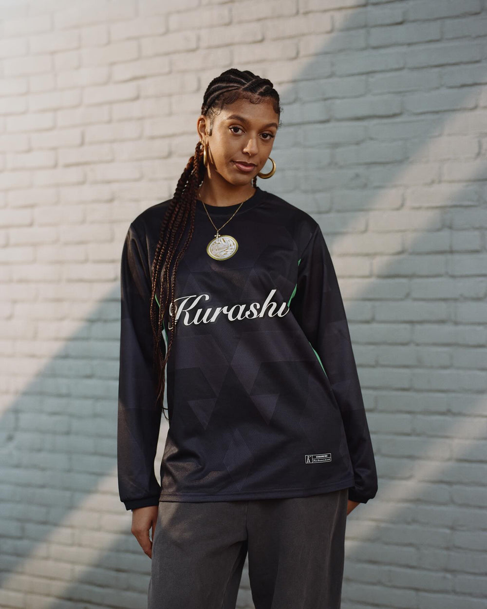 A young woman with long braided hair poses in front of a light gray brick wall, looking directly at the camera with a confident yet relaxed expression. She is wearing a black long-sleeve sports jersey with a subtle geometric pattern, green accents on the sides, and the word ‘Kurashi’ written in white cursive across the chest. A gold emblem is on the upper left side of the jersey. She accessorizes with large gold hoop earrings and a gold necklace with a circular pendant. The sunlight casts soft shadows on th