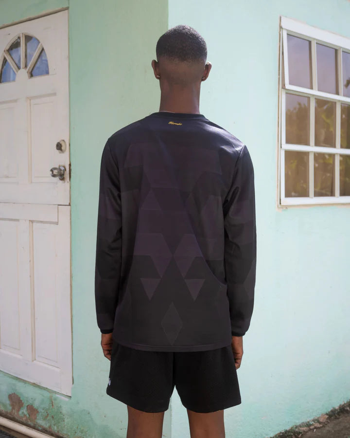 A young man with short hair stands with his back to the camera in front of a pastel green house with a white door and a window. He is wearing a black long-sleeve sports jersey with a subtle geometric pattern, and the word ‘Kurashi’ embroidered in small gold lettering at the top near the collar. He pairs the jersey with black athletic shorts. The setting is a simple residential area with a textured wall and natural lighting.