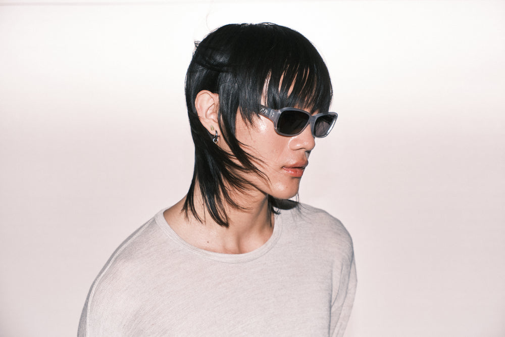 Person with stylish haircut and sunglasses wearing a light gray shirt on a plain background