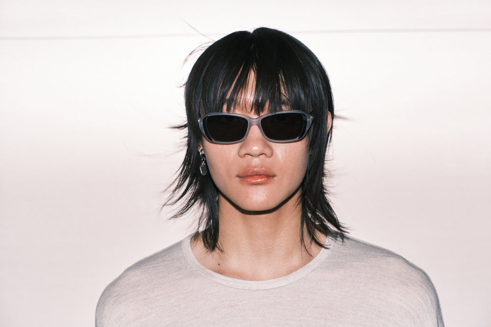 Model wearing rectangular black sunglasses with clear acetate frame, glossy nude lips, and sleek platinum blonde hair — minimalist fashion portrait on white background