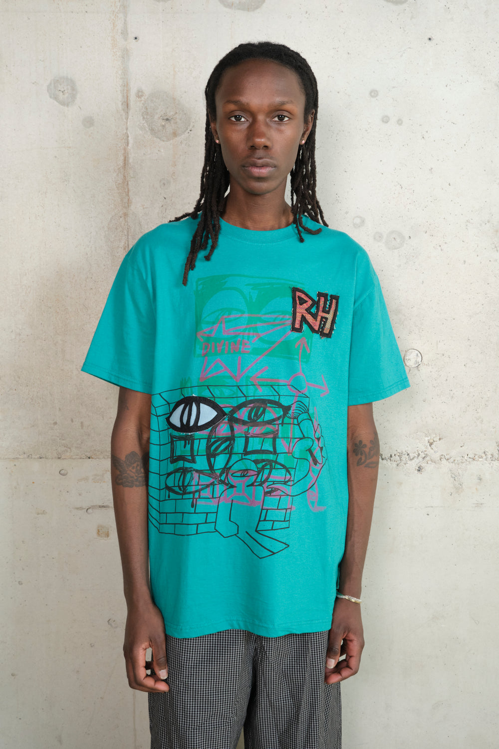 Model wearing turquoise streetwear T-shirt with abstract graffiti-style design, bold RH patch, and ‘DIVINE’ text, paired with loose-fitting checked trousers, standing against a concrete wall.