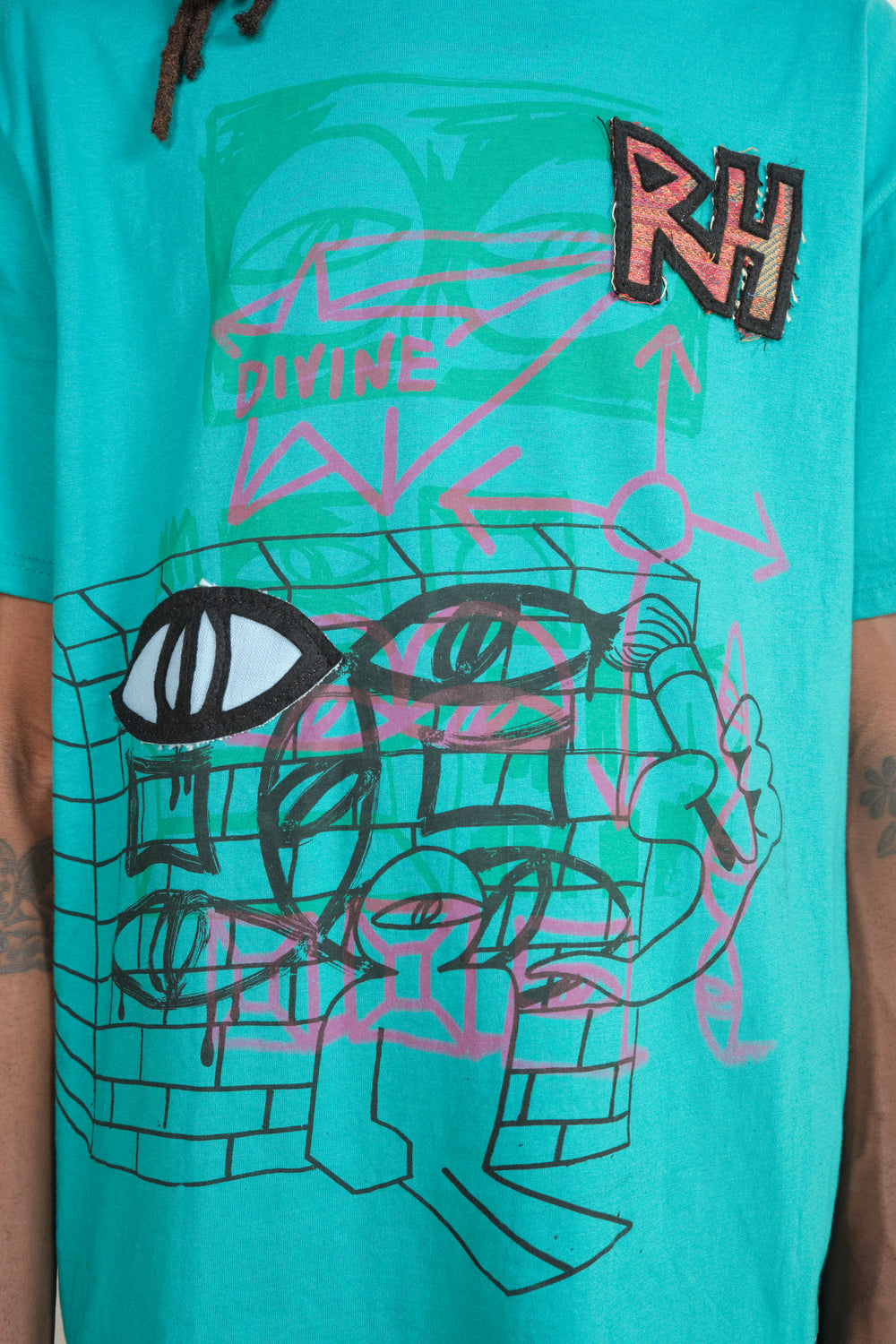 Close-up of turquoise T-shirt featuring abstract graffiti-style artwork with geometric shapes, layered eyes, arrows, the word ‘DIVINE’ in pink, and a textured ‘RH’ patch on the chest – streetwear graphic design detail.
