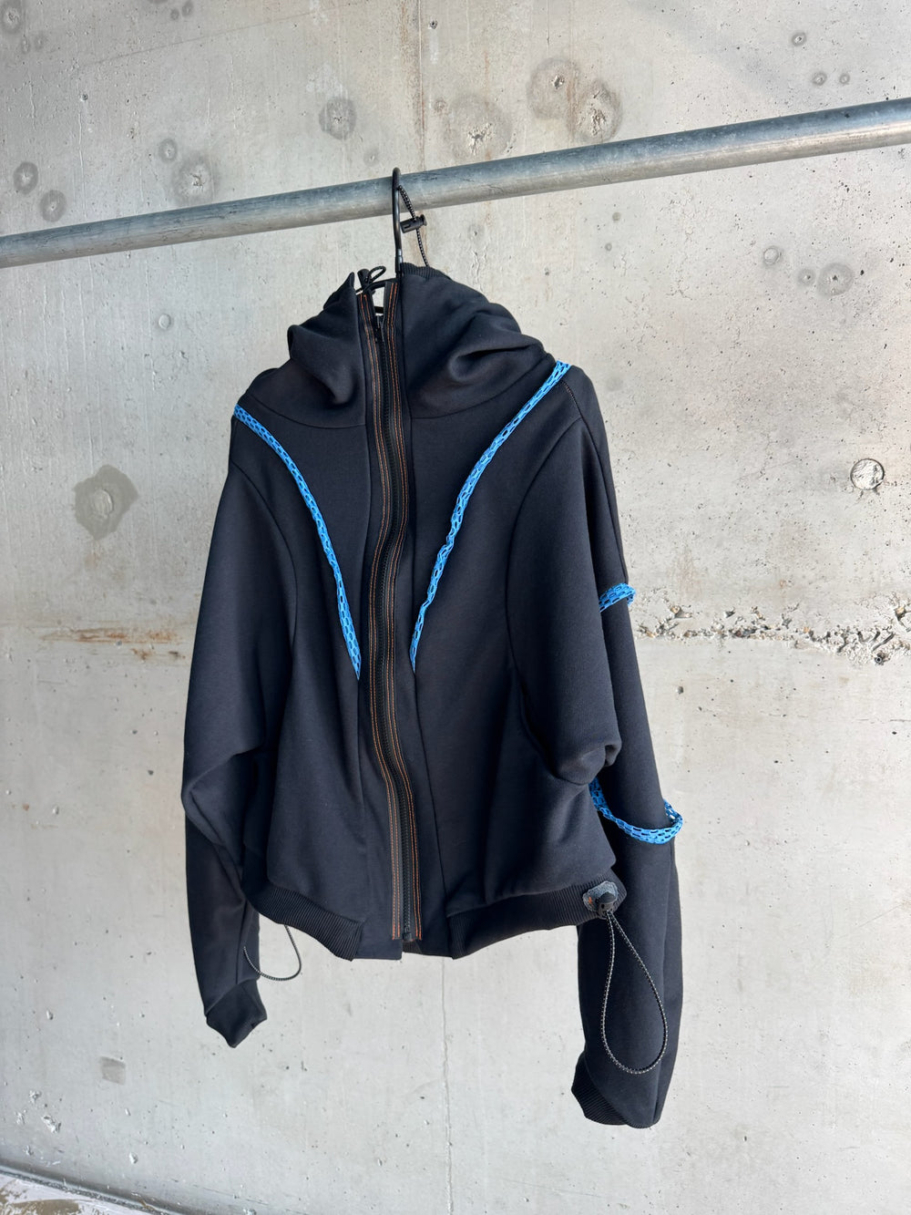Front view of a black zip-up hoodie with sculptural hood, blue decorative trim on shoulders and sleeves, orange contrast stitching, and adjustable drawstring details, displayed on a hanger against a concrete wall