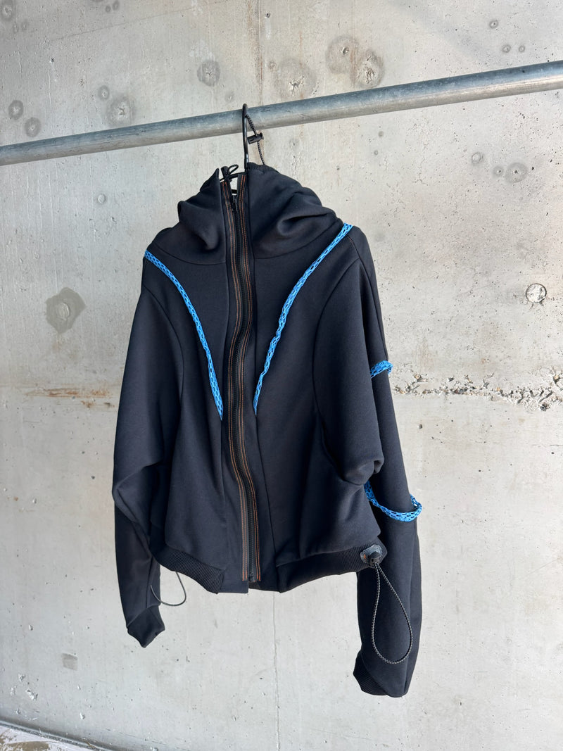 Front view of a black zip-up hoodie with sculptural hood, blue decorative trim on shoulders and sleeves, orange contrast stitching, and adjustable drawstring details, displayed on a hanger against a concrete wall