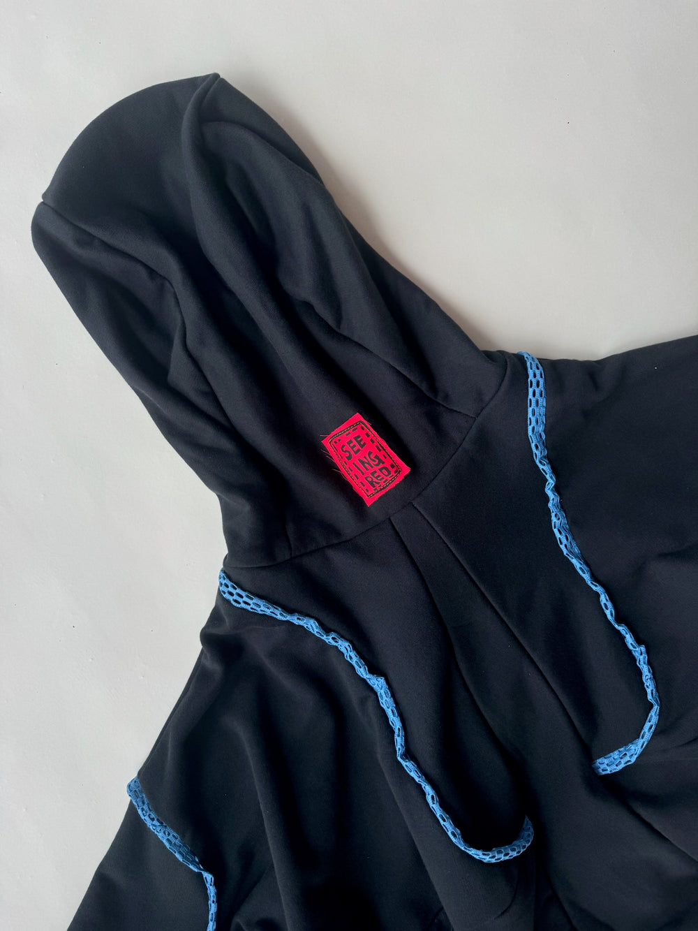 Close-up of a black hoodie with a bold red ‘SEEING RED’ label on the hood and intricate blue crochet detailing across the shoulders, highlighting handmade design elements