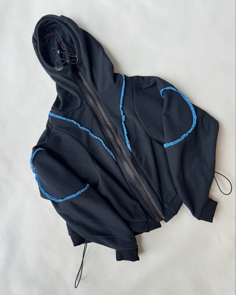 Flat lay of a black zip-up hoodie featuring blue decorative trim along the sleeves and shoulders, with a structured hood, orange contrast stitching, ribbed cuffs, and drawstring hem, displayed on a light background