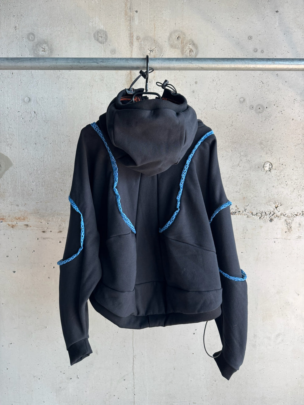 Back view of a black hoodie with sculptural hood, accented by blue decorative rope-like trim outlining the shoulders and sleeves, shown hanging against a textured concrete wall
