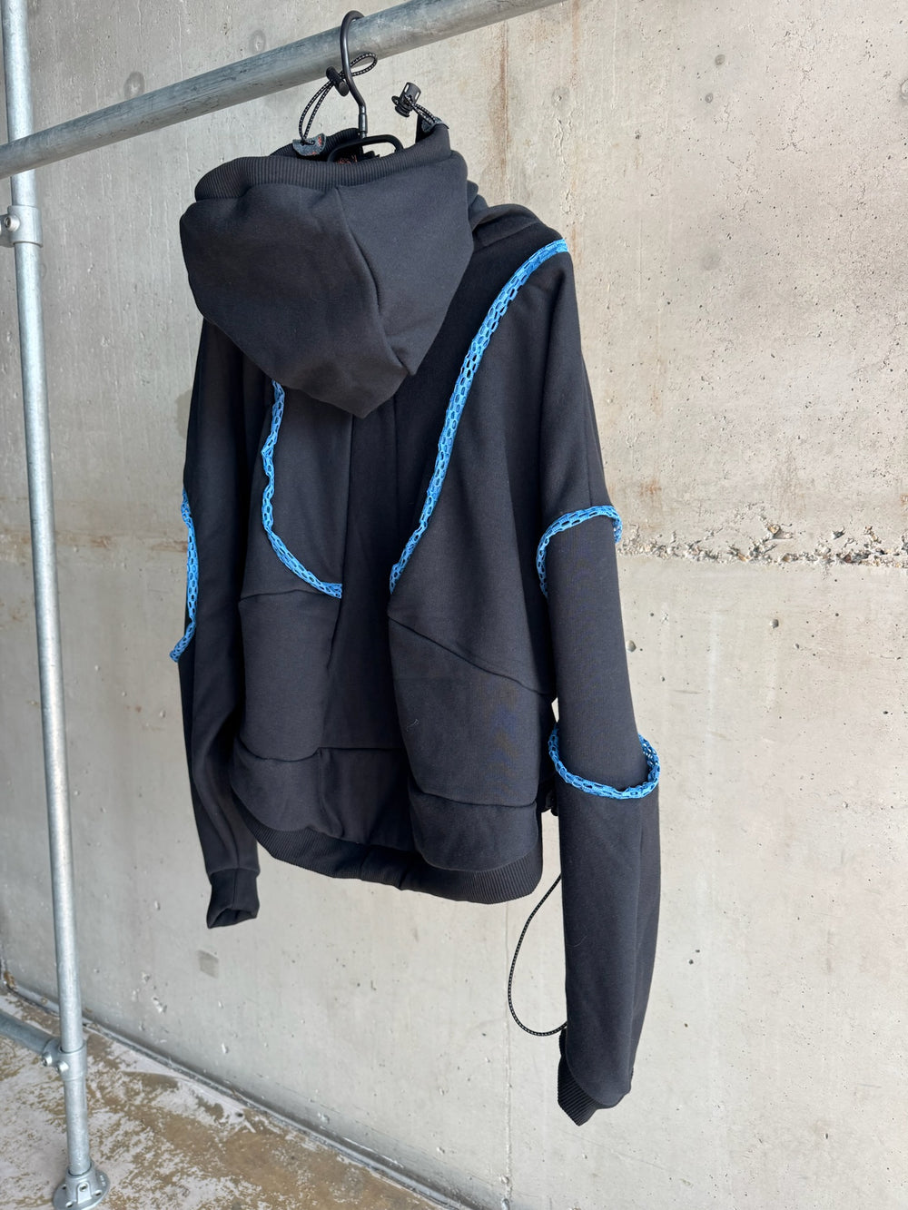 Side view of a modern black hoodie with sculptural design and bright blue braided trim details along the shoulders and sleeves, displayed on a hanger against a raw concrete wall