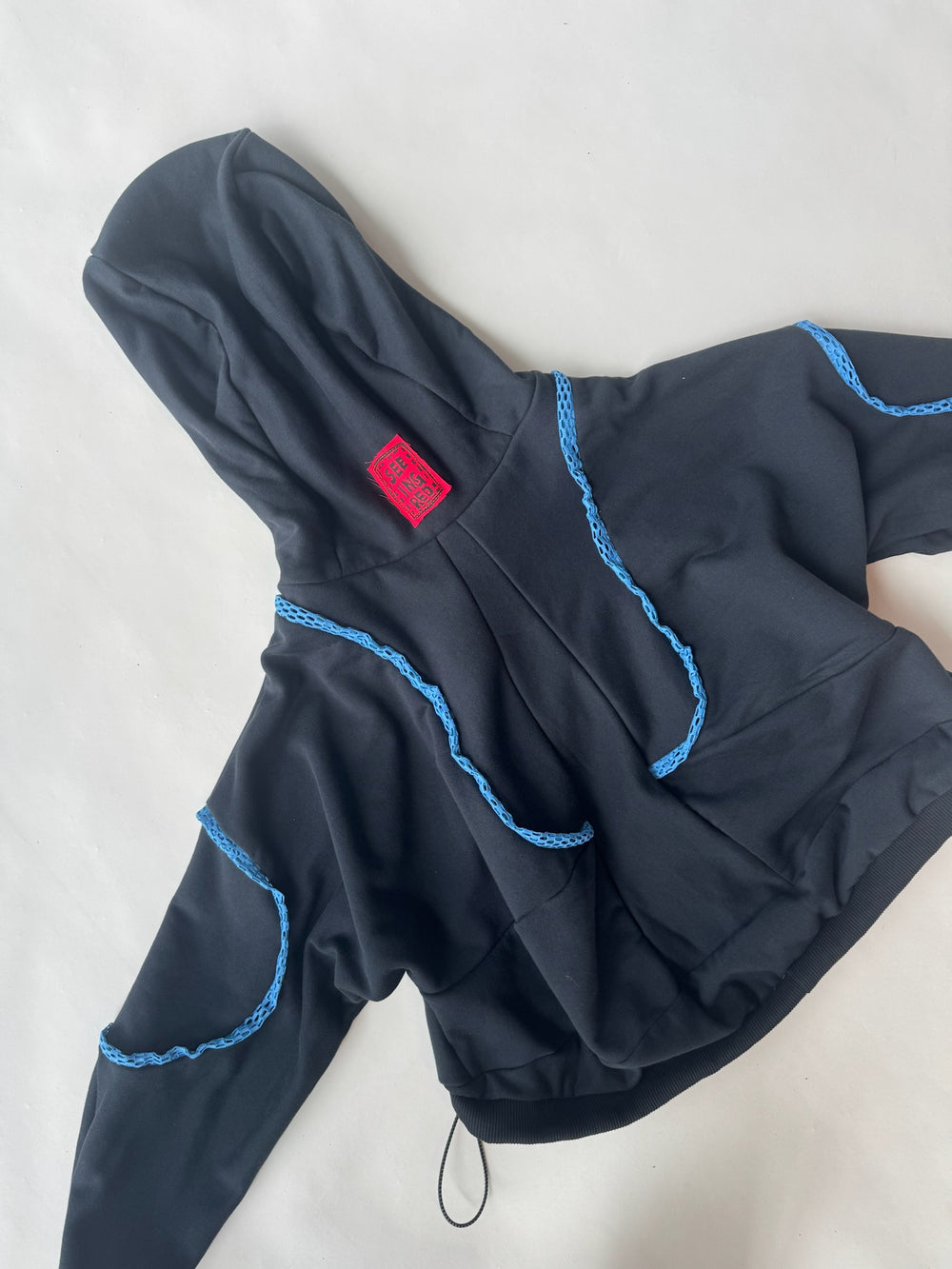 Back view of a black hoodie with blue crochet detailing and a red ‘SEEING RED’ label stitched on the hood, showcasing unique handcrafted embellishments and drawstring hem