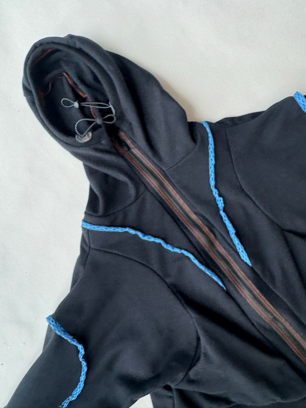 Close-up of a black hoodie with asymmetric zipper and blue crochet detailing along seams, highlighting the textured design and hood drawcords