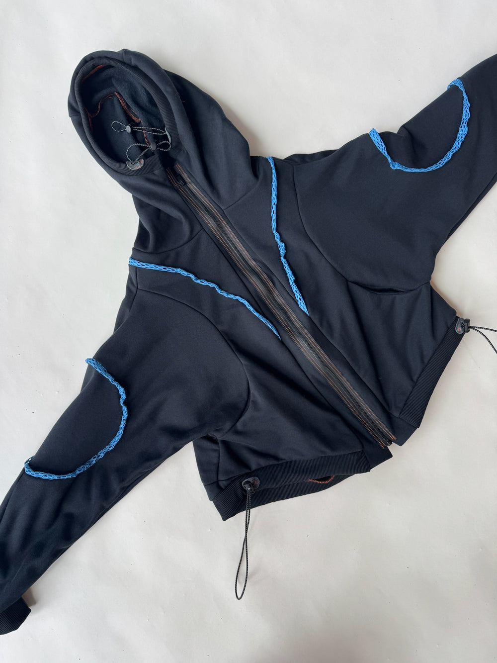 Flat lay of a black hoodie with asymmetric zipper, blue crochet-style seam accents, and adjustable drawcord hem, showcased on a light backgroun
