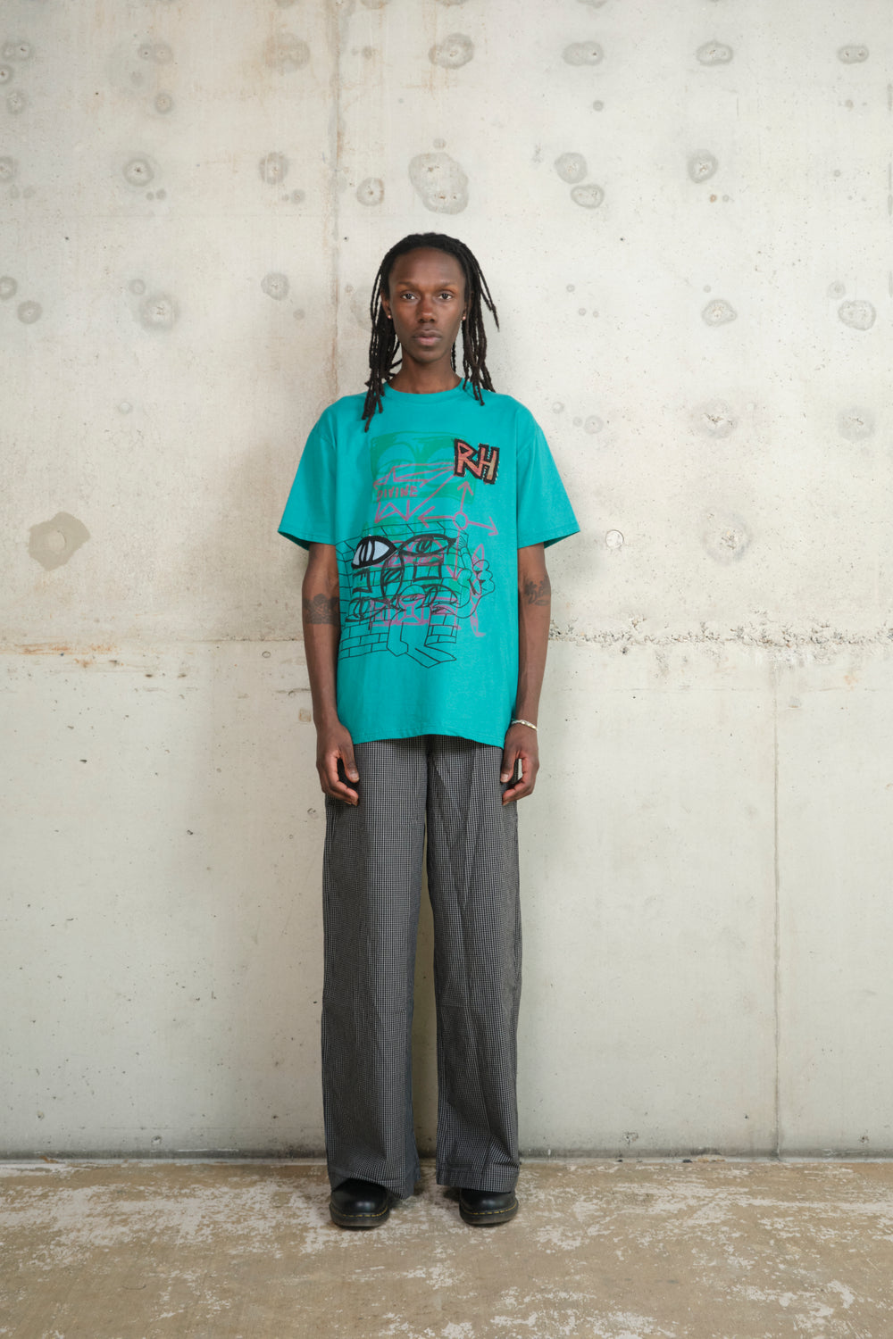 Model standing against a raw concrete wall, wearing a turquoise graphic T-shirt with abstract sketches, “RH” patch, and the word “DIVINE,” paired with black-and-white checkered wide-leg trousers and black shoes, blending playful streetwear with artistic flair.
