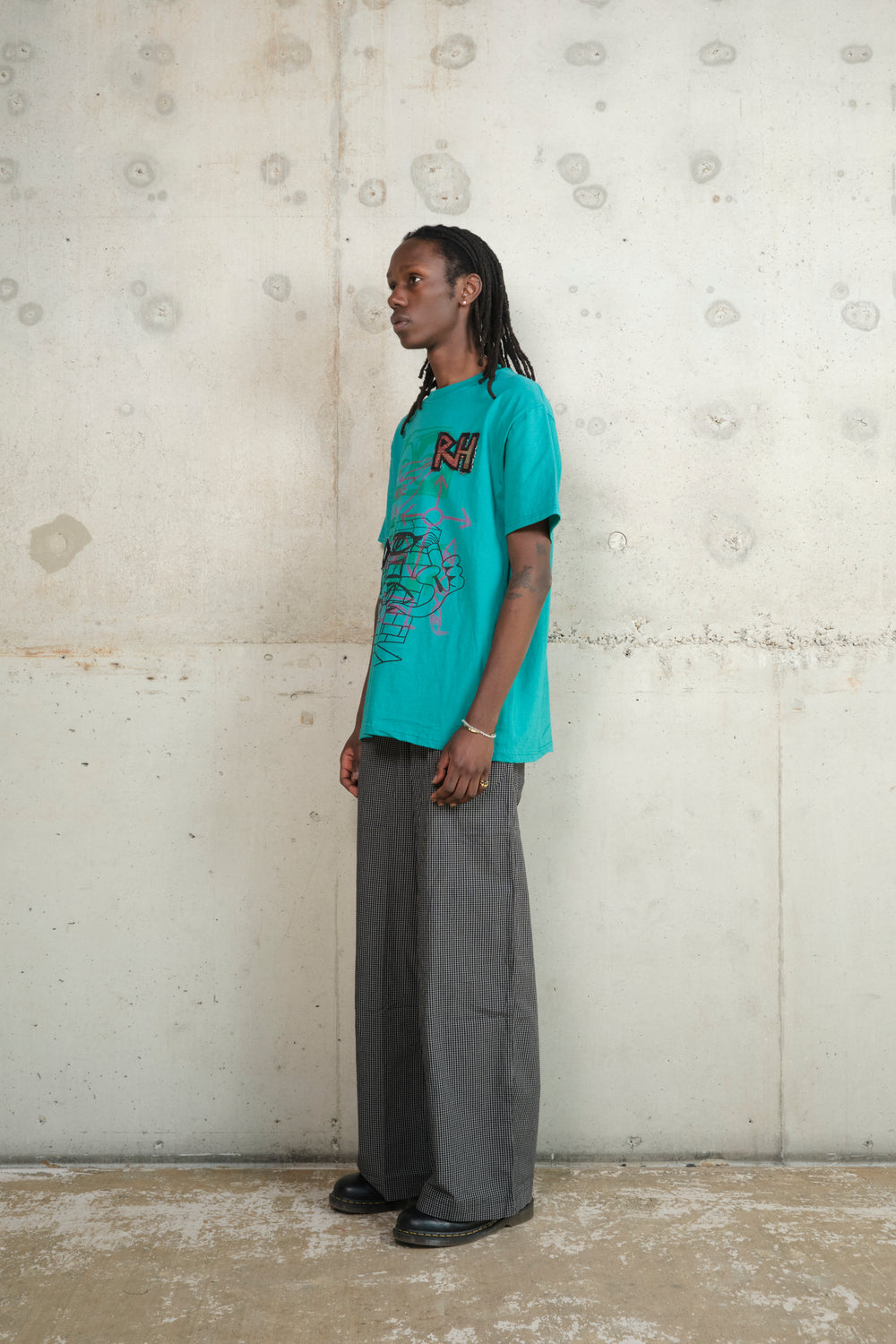 Model in a side pose against a raw concrete wall, wearing a turquoise graphic T-shirt with layered illustrations and an “RH” patch, styled with loose-fitting checkered trousers and black shoes, merging retro graphics with relaxed tailoring.