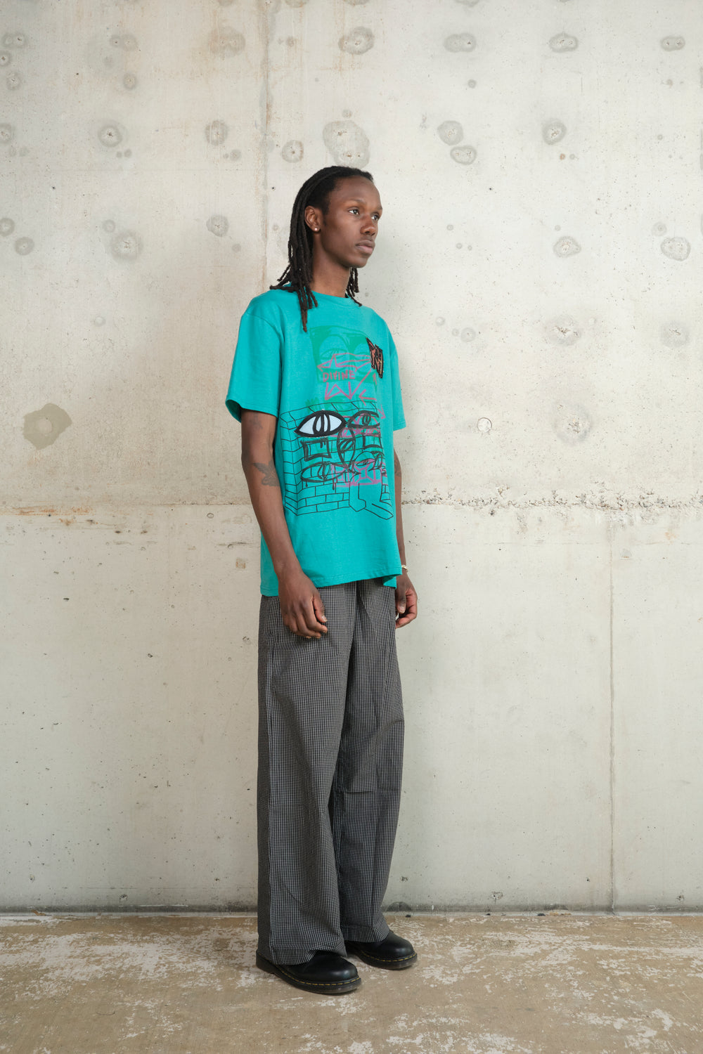 Model wearing turquoise graphic T-shirt with abstract graffiti-style illustration and ‘RH’ patch, paired with black-and-white checkered wide-leg trousers and black shoes, standing against a raw concrete wall – urban streetwear fashion editorial