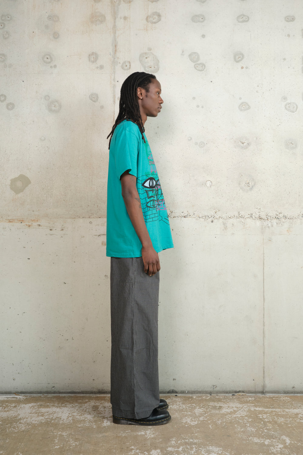 Model standing in profile against a concrete wall, wearing a turquoise graphic T-shirt featuring a bold “RH” patch and abstract multicolor print, paired with loose-fitting black and white checkered trousers and black shoes — combining streetwear edge with casual sophistication.
