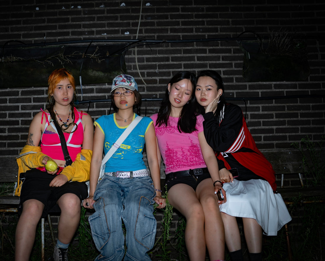 Four friends sit on a wooden bench against a brick wall, dressed casually in colorful, streetwear-inspired outfits. One person holds a green apple, while the others pose with a relaxed, laid-back attitude, conveying a sense of togetherness and self-expression.
