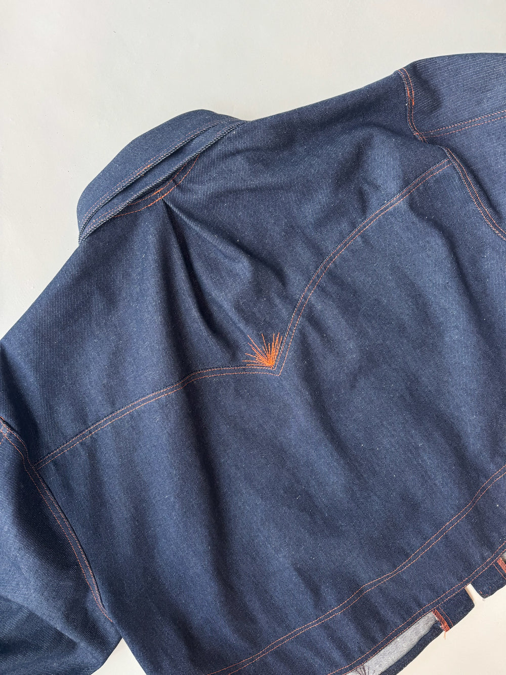 Back detail of a dark indigo denim jacket featuring orange contrast stitching and a unique sunburst embroidery accent below the shoulder seam, laid flat against a neutral background.