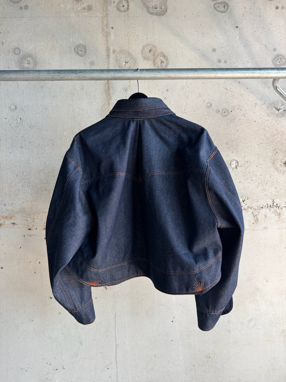 Back view of a dark indigo denim jacket featuring orange contrast stitching and structured tailoring, displayed on a hanger against a raw concrete wall backdrop.