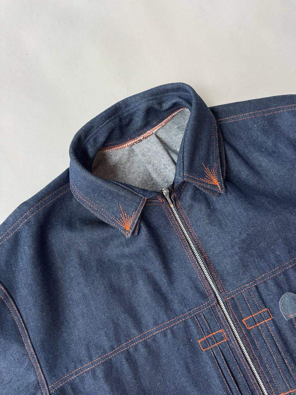 lose-up of the collar and upper chest of a dark indigo denim jacket with bright orange contrast stitching and a silver zipper, displayed against a neutral light background.