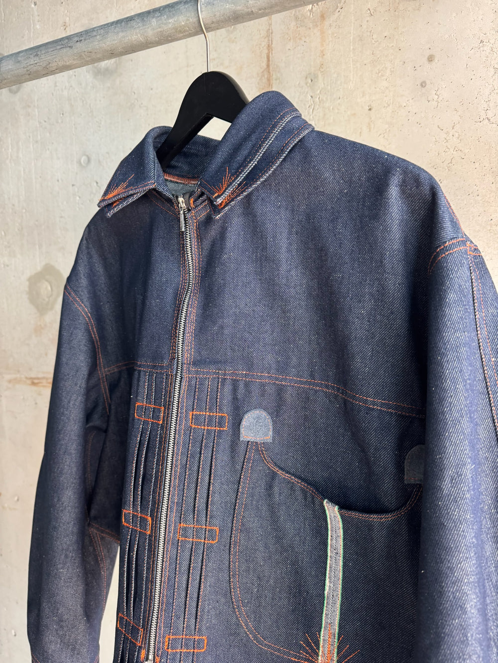 Close-up of avant-garde indigo denim jacket with contrasting orange stitching, sculptural seams, and curved pocket detail, hanging against a raw concrete wall.