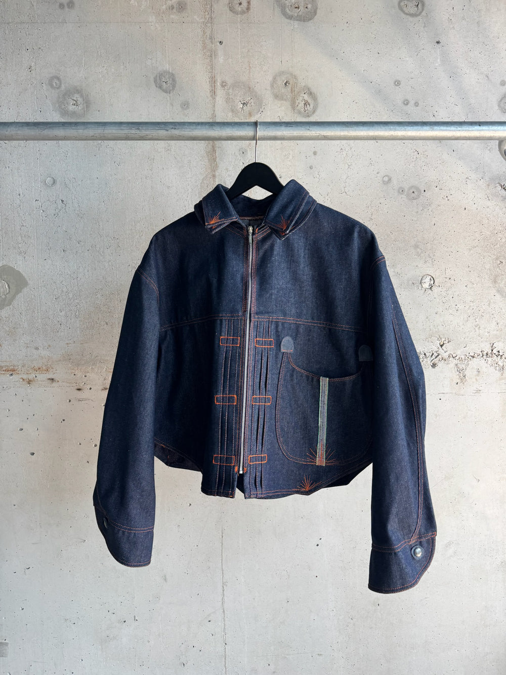Dark indigo denim jacket with asymmetrical orange contrast stitching, zip-up front, and curved detailing on the pockets, photographed against a raw concrete wall on a black hanger.