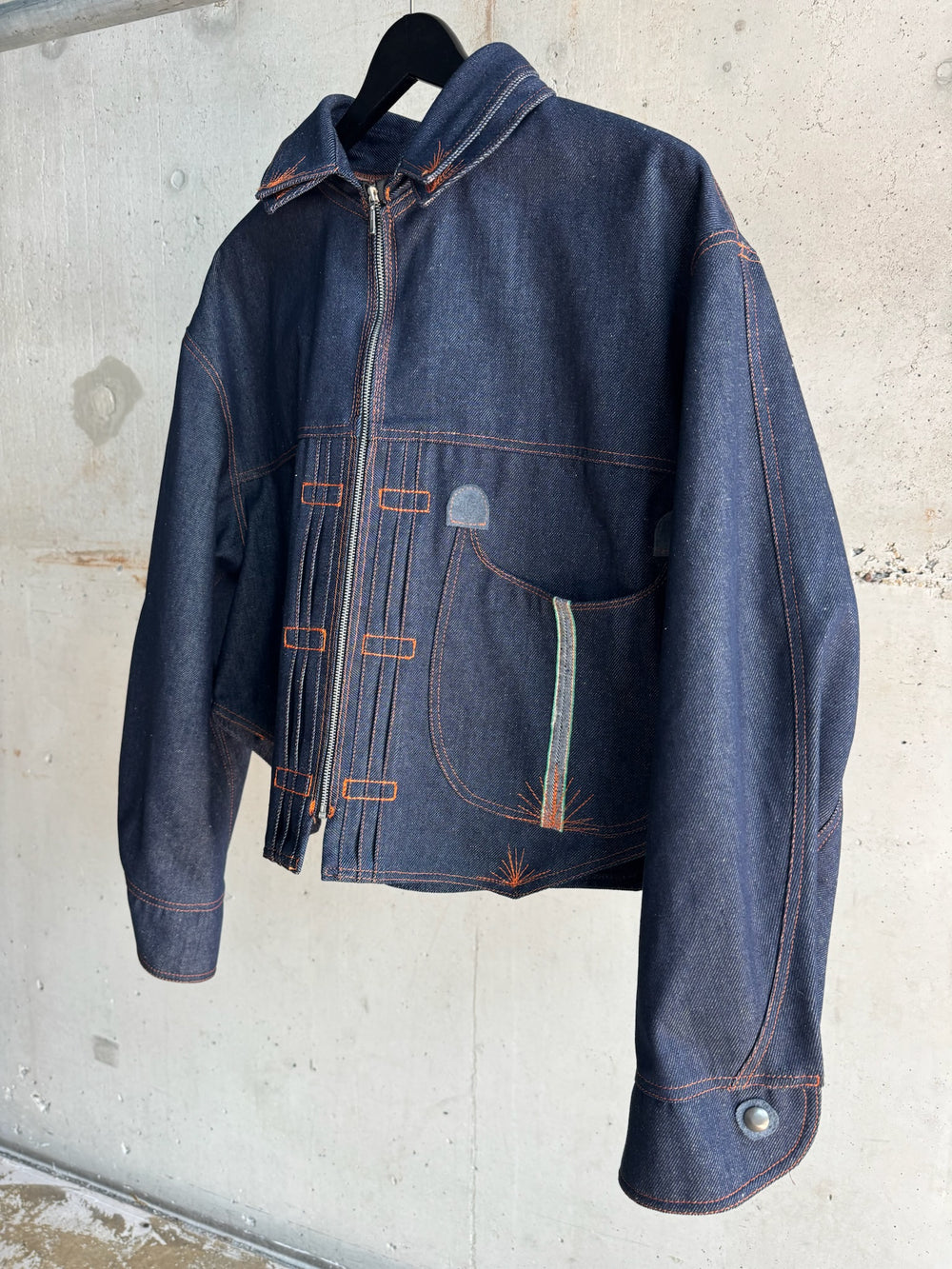 Contemporary cropped denim jacket in deep indigo with asymmetric zipper, orange topstitching, and sculptural curved pocket, displayed on hanger against industrial concrete backdrop.