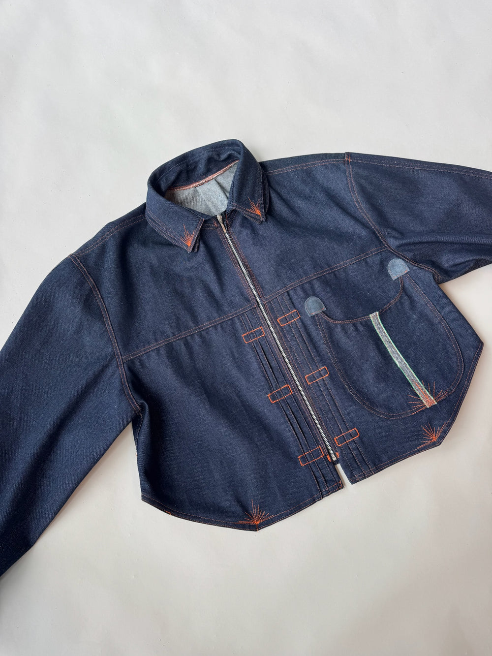 Flat lay of a dark indigo denim jacket with asymmetrical orange topstitching and a curved front pocket featuring a mint green accent, set against a light grey background.