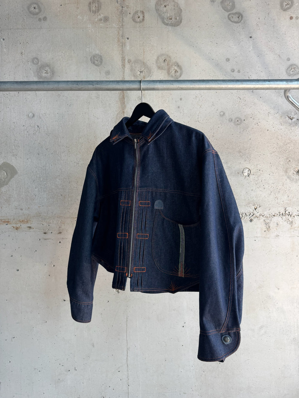 Dark indigo denim jacket with asymmetrical orange contrast stitching, zip-up front, and curved detailing on the pockets, photographed against a raw concrete wall on a black hanger.