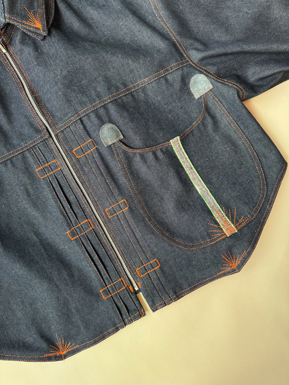 Close-up of a dark indigo denim jacket showcasing detailed orange topstitching, a rounded front pocket with multicolored embroidery, and a vertical zipper closure with geometric stitched accents on a light beige background.