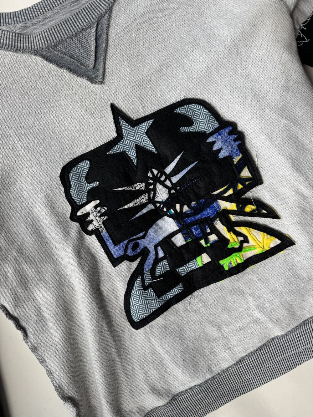 Close-up view of grey sweatshirt showcasing bold abstract skull patch with layered fabric textures in black, blue, yellow, and geometric patterns, emphasizing the intricate design and distressed hem